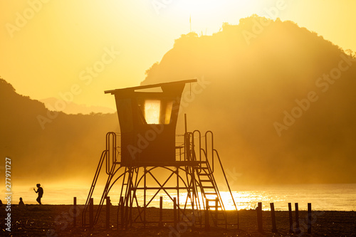 Silhouette of an old lifeguard tower against a glowing haze on the beach of Copacabana in Rio de Janeiro backlit by intense golden hour sunlight with a mountain in the background