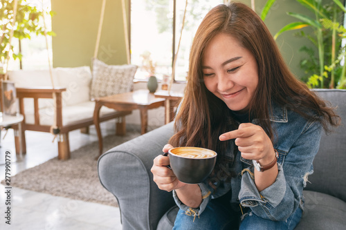 Closeup image of a happy beautiful asian woman holding and pointing finger at a cup of hot coffee in cafe