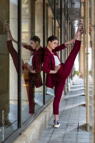 beautiful girl with short hair brunette in sunglasses shows stretching near the glass wall of the business center. The gymnast trains in the urban environment. Women's business suit in red.