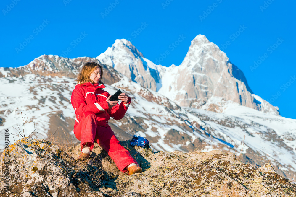 A woman in a red jacket with a phone sits in the mountains. A woman in a red jacket with a phone sits in the mountains.