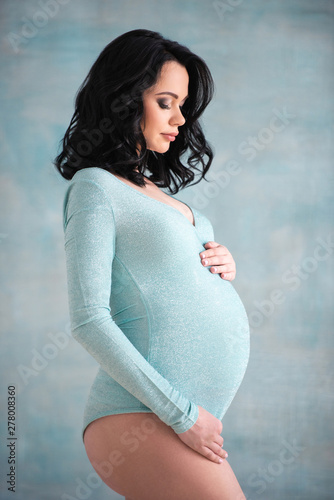 Beautiful pregnant girl brunette woman in a trendy blue body suit stands near the wall. Concept of healthy pregnancy and happy motherhood