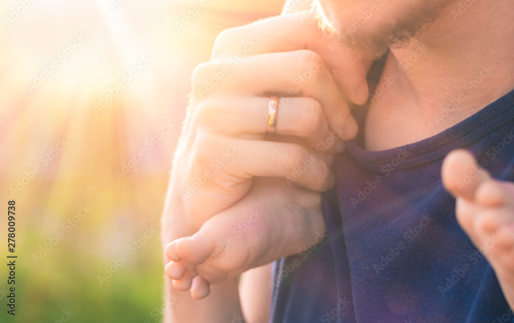 Naklejka premium Man's hand with a wedding ring on his finger, holding the foot of his bare little child. Toddler is sitting at his father's neck on the background of soft sunlight and blurred green bokeh.