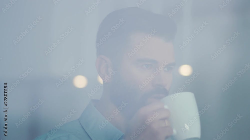 Young businessman drinking coffee and admire view standing by window in office attractive boss enjoying tea caucasian looking young break manager executive observing relaxation view slow motion Young businessman drinking coffee and admire view standing by window in office attractive boss enjoying tea caucasian looking young break manager executive observing relaxation view slow motion
