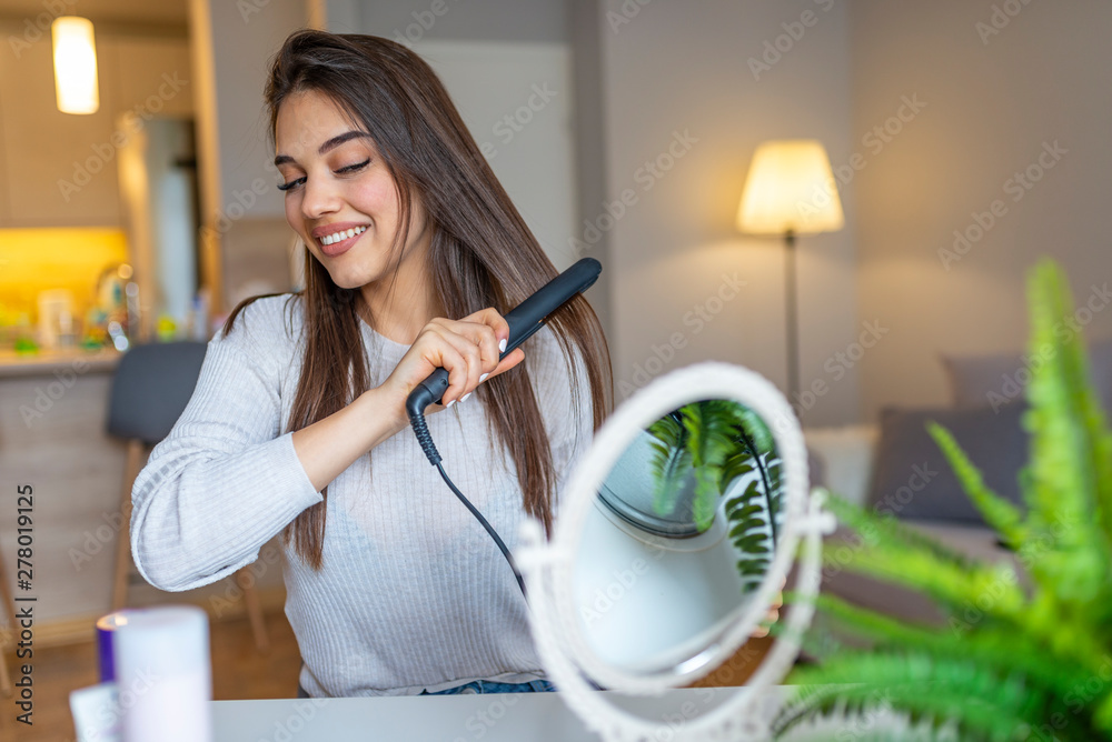 Beautiful girl is using a hair straightener and smiling while looking into  the mirror at home. Smiling woman straightening hair with hair straightener  at home. Stock Photo | Adobe Stock