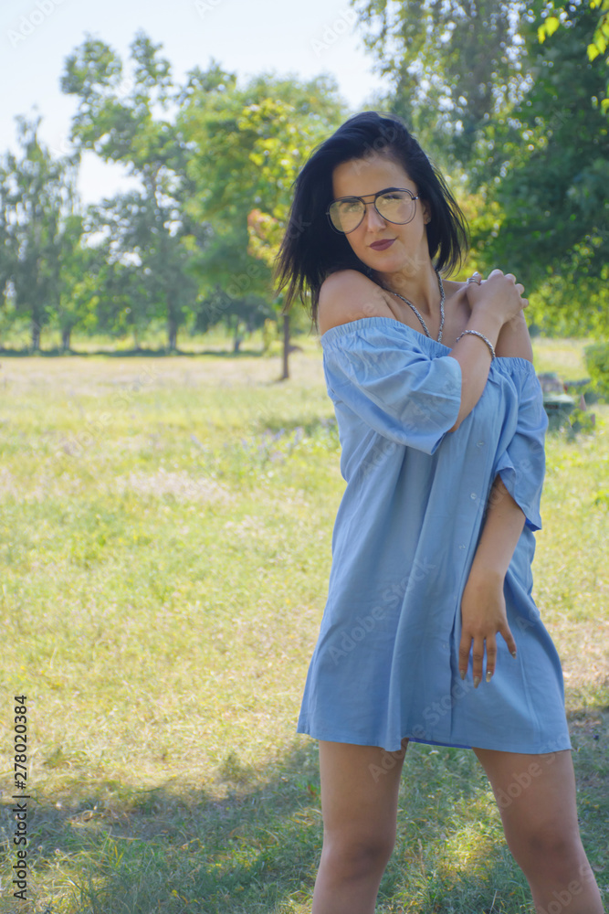 Beautiful young brunette woman wearing blue dress, smiling, walking in the park, is happy. Minimalist clothes. Human natural beauty. Trendy casual summer or spring outfit. Street fashion. Beautiful young brunette woman wearing blue dress, smiling, walking in the park, is happy. Minimalist clothes. Human natural beauty. Trendy casual summer or spring outfit. Street fashion.