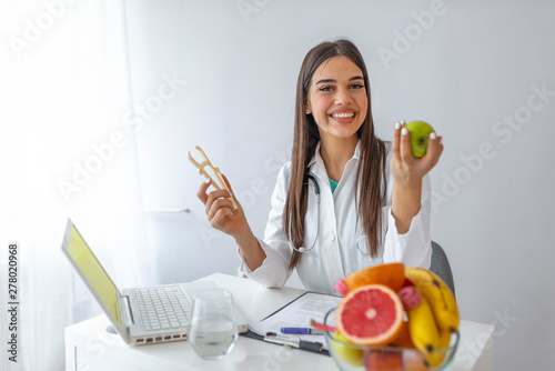 Young female nutritionist sitting at table with fresh vegetables and fruits in her office. Female nutritionist sitting at table with clipboard and healthy products on white background photo
