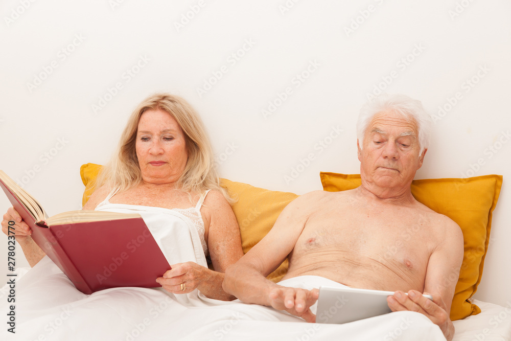 COUPLE SENIOR LISANT LIVRE ET TABLETTE DANS UN LIT COUPLE SENIOR LISANT LIVRE ET TABLETTE DANS UN LIT