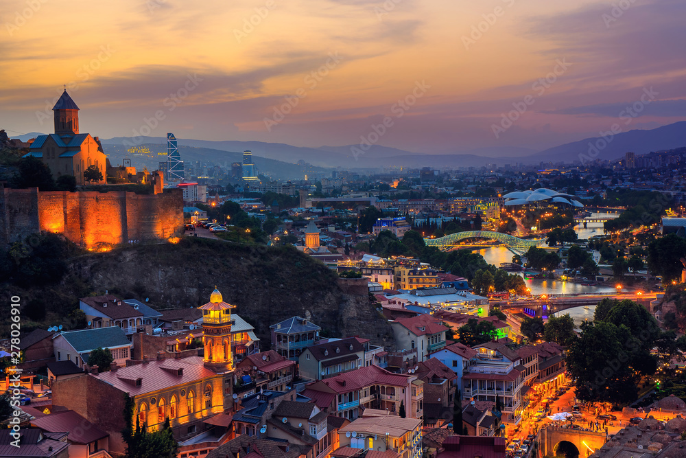 Beautiful view of the Georgian capital Tbilisi at sunset Narikala fortress in the foreground, Kura river with a bridge and mountains in the background.