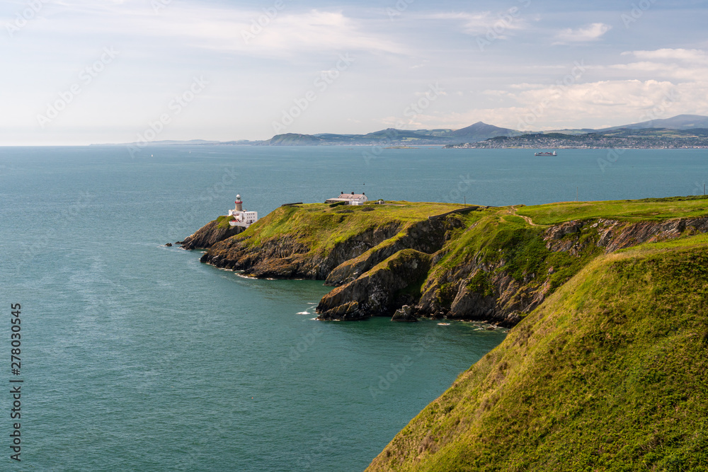 Scenic view over the Irish Coast with sheer cliffs covered in green vegetation and the Baily Lighthouse in the distance. Landscape from the Howth Cliff Walk in Dublin, Ireland. Scenic view over the Irish Coast with sheer cliffs covered in green vegetation and the Baily Lighthouse in the distance. Landscape from the Howth Cliff Walk in Dublin, Ireland.