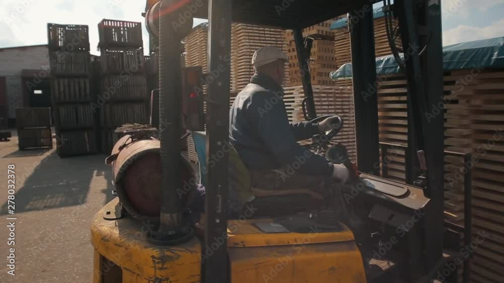 Forklift operator handling wooden pallets in warehouse. Man using loader dor Pack of wooden planks.