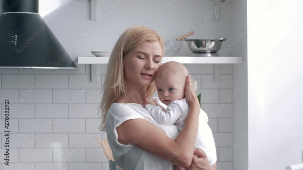 loving mother holds on hands newborn baby girl, portrait of young woman gently hugs and kissing her child at kitchen close up