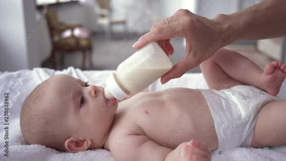 mother care, baby girl in diaper is lying on the changing table and drinking milk from a bottle that mom holds in her hands close-up
