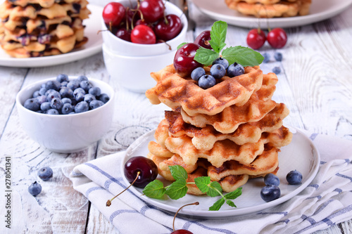 Stack of waffles topped with fresh bilberries, cherries and mint on the white wooden table