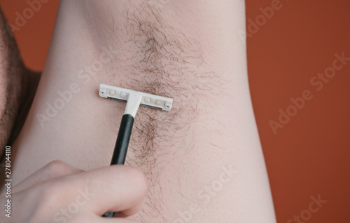 Attractive man with a blue razor in his hands is going to shave hair with armpits isolated on orange background