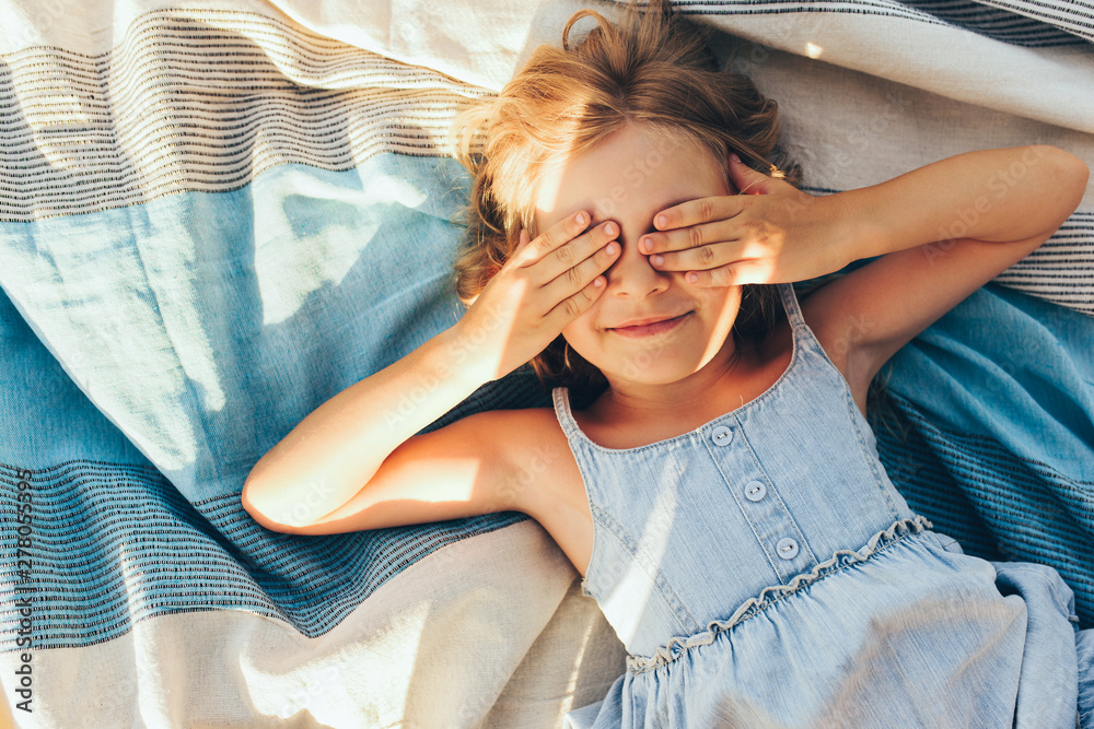 Beautiful blonde little girl lying on the blanket, cover her eyes from the sun with both hands, enjoying summer day. Adorable child having fun and plaiyng peekaboo outdoors in the park - obrazy, fototapety, plakaty 