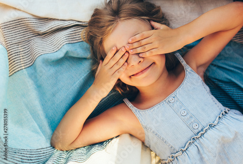 Принти на полотні Beautiful blonde little girl lying on the blanket, cover her eyes from the sun with both hands, enjoying summer day