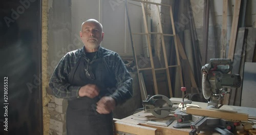 Senior gray haired male carpenter with beard standing at wood manufacture with crossed arms smiling positively into camera. photo