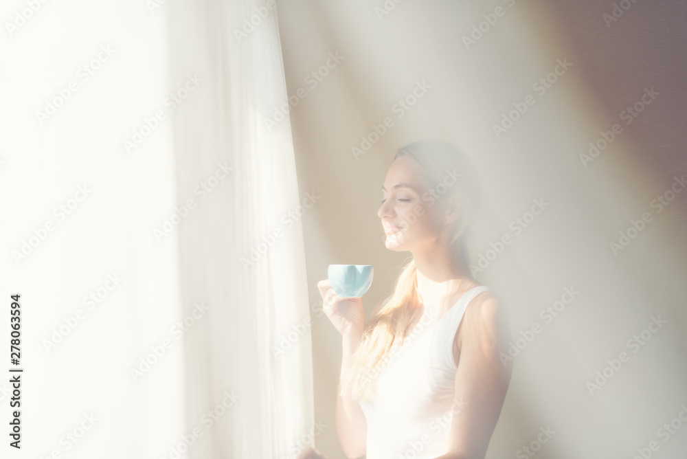 Young beautiful women drinking coffee in the mornng bedroom near window with white curtain overlay. Happy caucacian girl having a good time drinking tea.