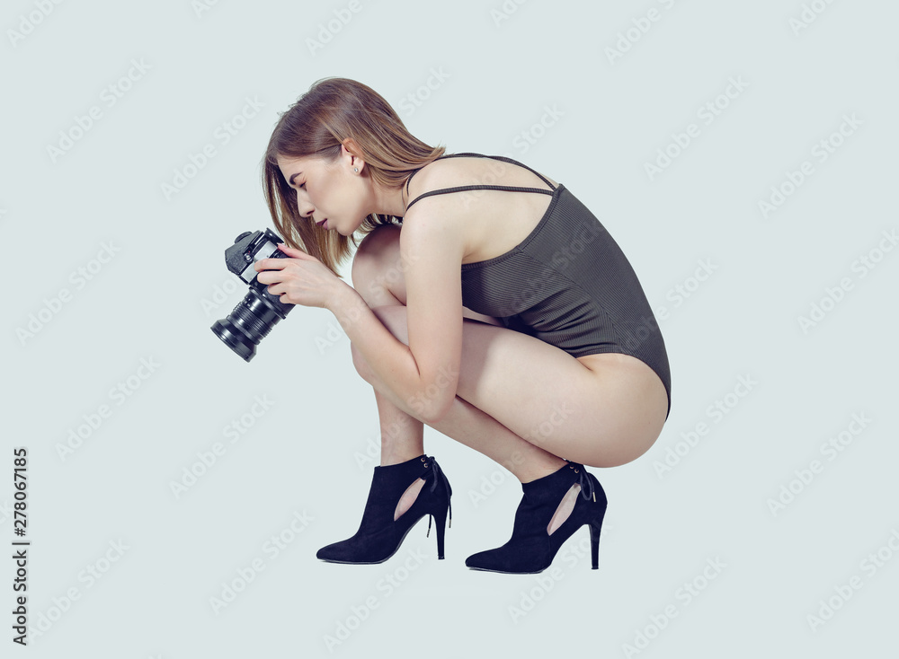 Sexy girl with a short haircut in a dark bodysuit in black boots on a high heel is squatting, holding a vintage film camera in her hands in the studio on a light background. Isolate