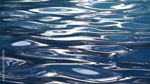 Water Surface Background stock footage features an extreme close-up shot of blue water in a swimming pool. The water is rippling in slow motion and has some reflections of the sun.