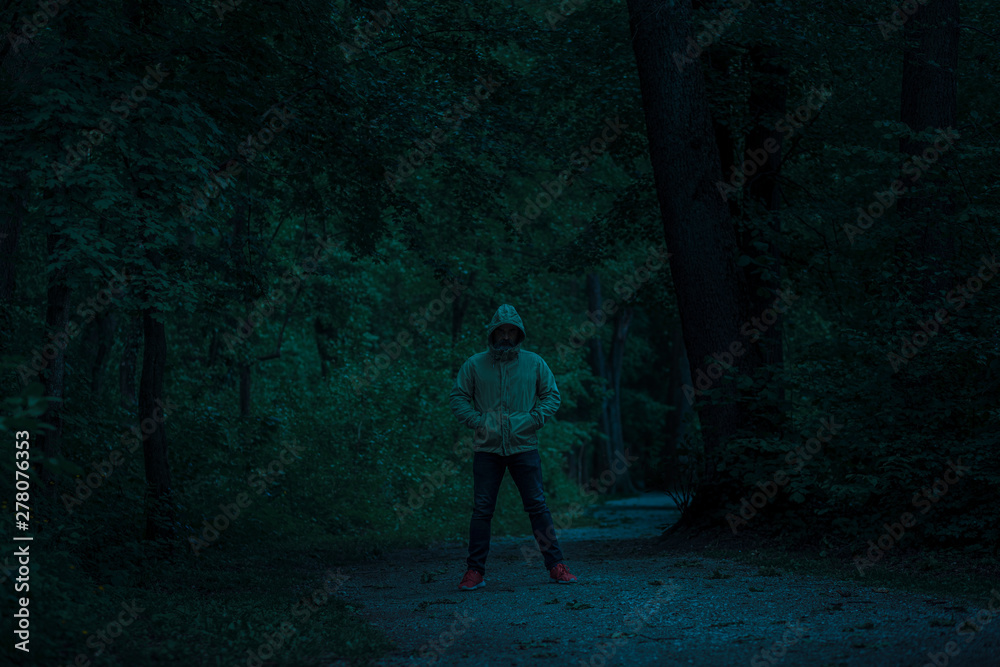 Scary dark male figure in a hoodie standing on the path in the forest with mysterious light coming behind him, night horror scene in forest