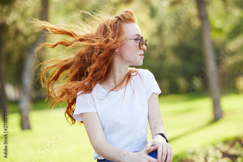 Portrait of a beautiful red-haired girl who sits in the park and looks away. The wind develops her hair and the girl smiles