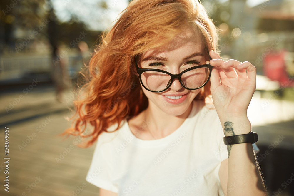 Portraits of a charming red-haired girl with a cute face. Girl posing for the camera in the city center. She has a wonderful mood and a lovely smile - obrazy, fototapety, plakaty 
