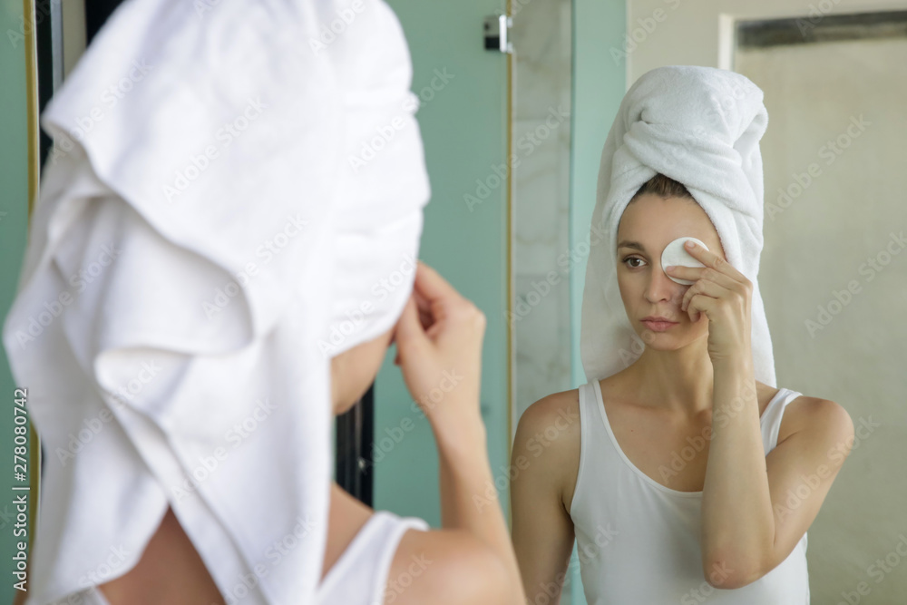 Fototapeta premium Portrait of a beautiful young woman with a towel on the head standing in the bathroom and cleaning her face with cotton pads in front of the mirror, removing makeup, beauty and spa concept