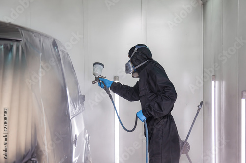 A male worker in jumpsuit and blue gloves paints with a spray gun a side part of the car body in black after being damaged at an accident. Auto service industry professions