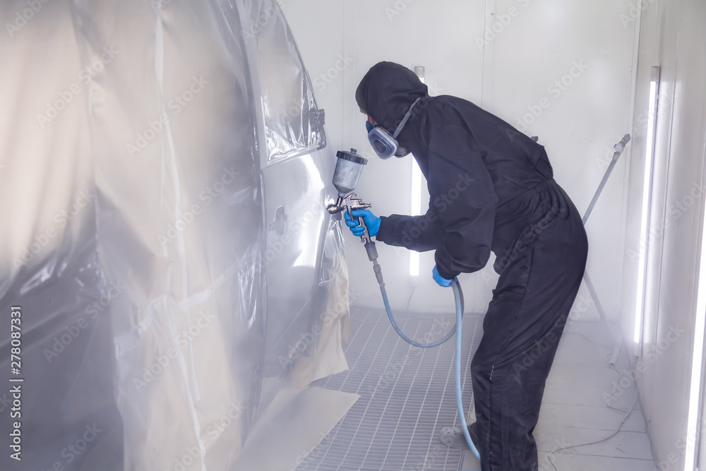 A male worker in jumpsuit and blue gloves paints with a spray gun a side part of the car body in black after being damaged at an accident. Auto service industry professions