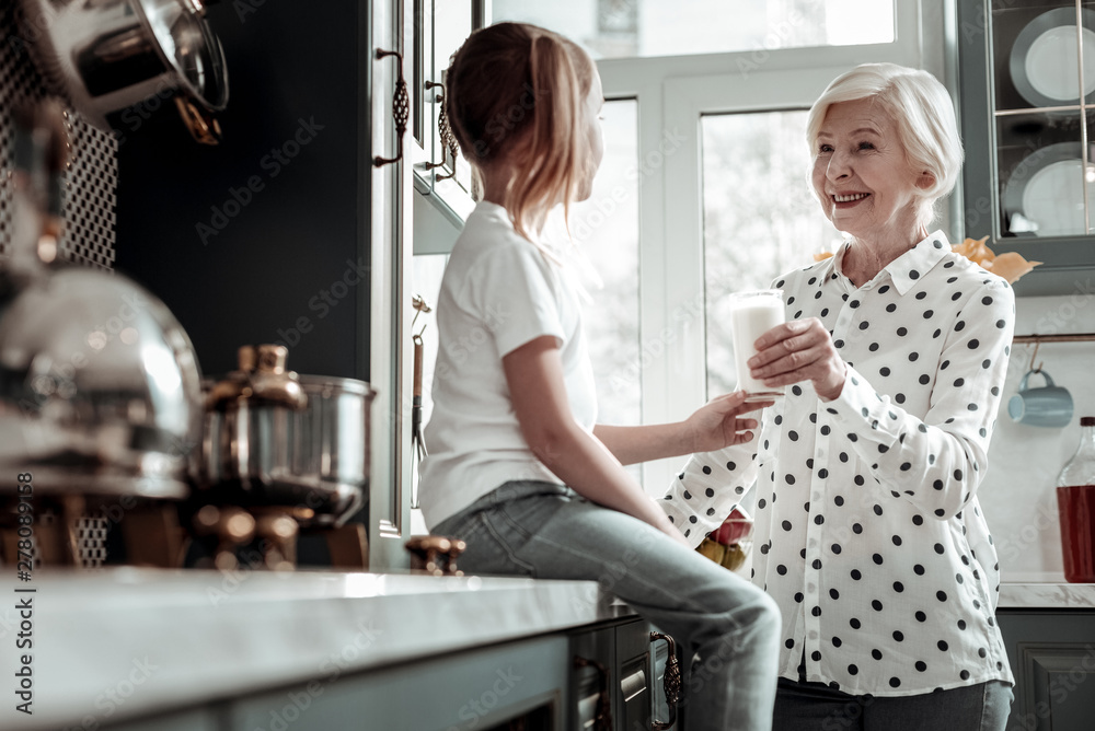 Kind loving grandmother standing in the kitchen and offering milk to girl Kind loving grandmother standing in the kitchen and offering milk to girl
