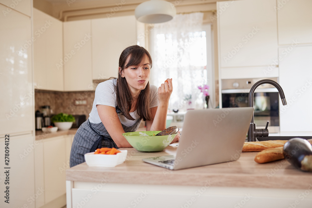 Gorgeous Caucasian brunette in apron leaning on kitchen counter, looking at laptop, looking for new recipe and eating carrot. On table is lots of vegetables. Gorgeous Caucasian brunette in apron leaning on kitchen counter, looking at laptop, looking for new recipe and eating carrot. On table is lots of vegetables.