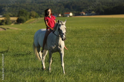 Teen girl ride on white horse without saddle on meadow in late afternoon, sunset time