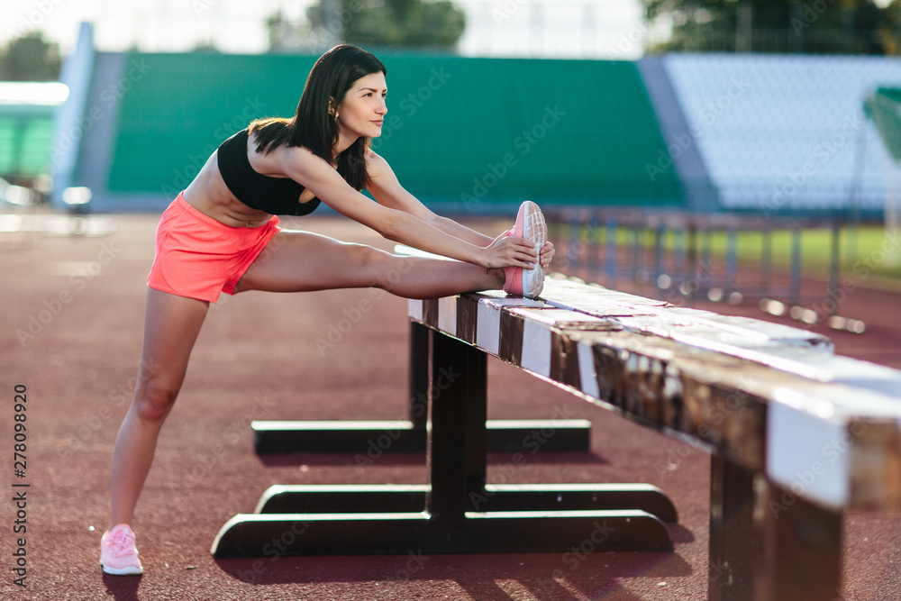 Beautiful brunette runner woman doing stretching leaning her leg on barrier for running stretching before workout - outdoor shot. Sport girl doing exercise in the stadium