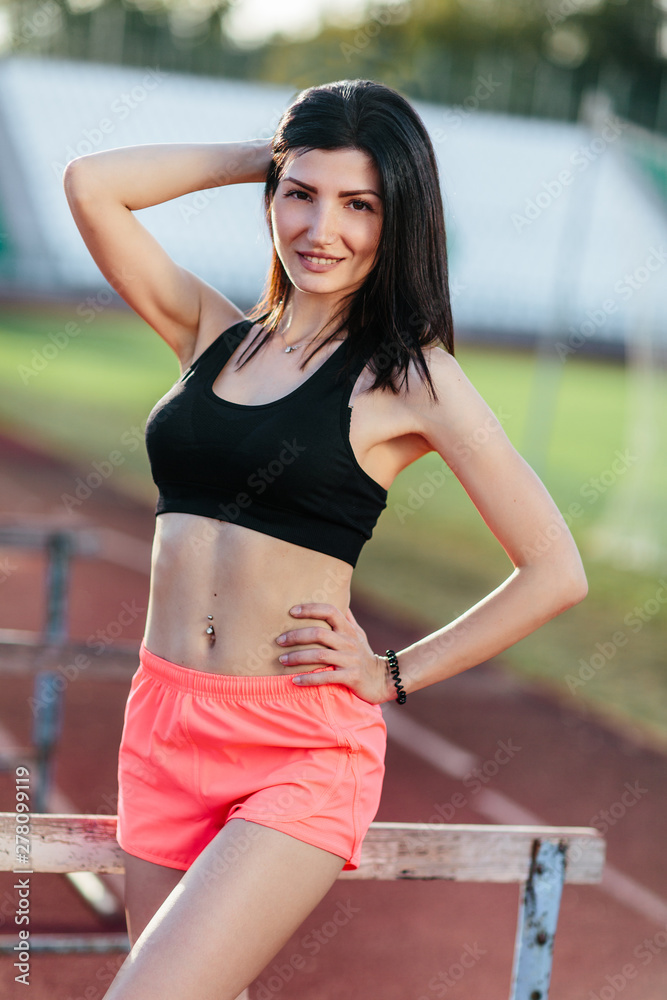 Portrait of young brunette woman athlete on stadium sporty lifestyle standing on track posing near the barriers running jumping to camera smiling playful