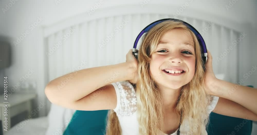 Adorable teenage girl sitting on bed in headphones, listening to music and having fun, relaxation after school day