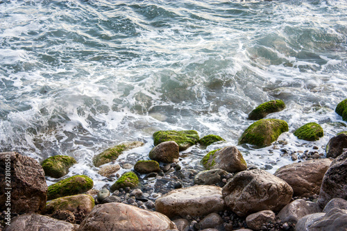 Stone beach of the Black Sea with magnificent waves of the surf. View of the surf waves on the sea beach on a summer day. Foam from the surf on the stones close-up.