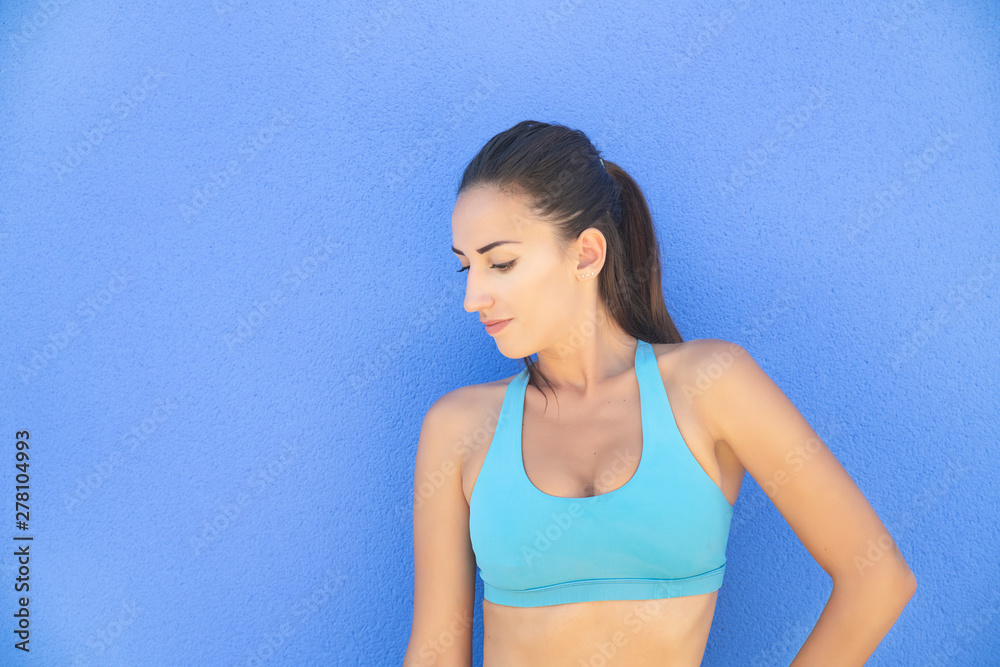 Fit Caucasian woman wearing a blue sports bra and leaning against a blue background, posing with a pensive, relaxed look after an intense workout routine, healthy lifestyle, sport or fitness concept Fit Caucasian woman wearing a blue sports bra and leaning against a blue background, posing with a pensive, relaxed look after an intense workout routine, healthy lifestyle, sport or fitness concept