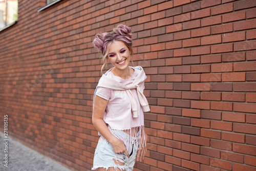 Portrait of a young beautiful girl with pink hair bun smiling against a red brick wall.