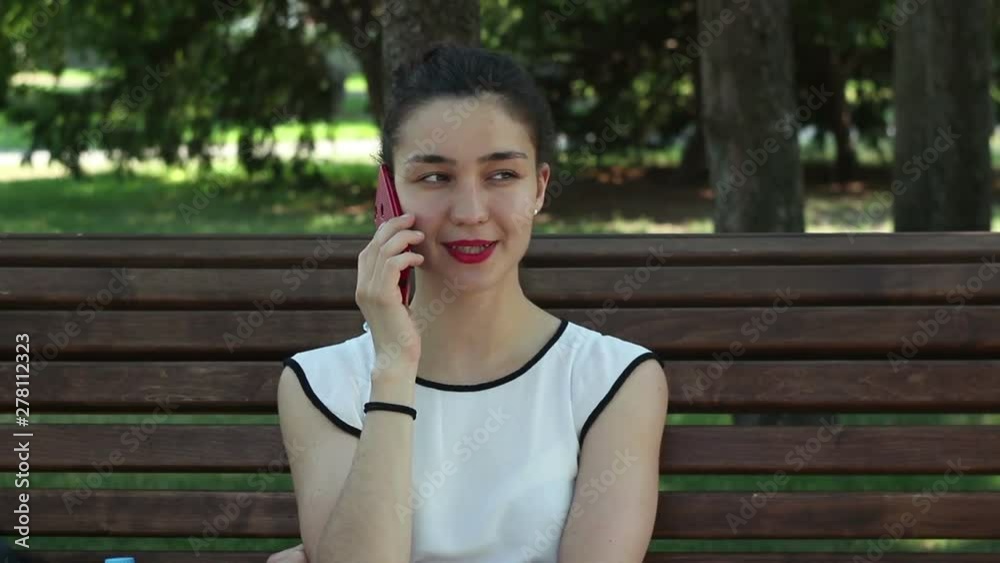 Young beautiful girl talking on the phone and smiling sitting on a bench in the park. Young beautiful girl talking on the phone and smiling sitting on a bench in the park.