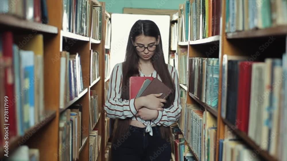Beautiful Girl with long black hair looks straight into the camera and walks along beetwen the shelves in the library and holds books in hand. Slow motion Beautiful Girl with long black hair looks straight into the camera and walks along beetwen the shelves in the library and holds books in hand. Slow motion