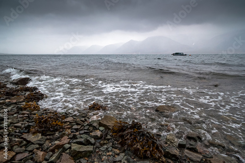 Sea bay in the rain. Severe seascape with coast and mountains. Boat in the sea. Surf and seaweed. Autumn weather. Arctic nature. Egvekinot Bay, Kresta Gulf, Bering Sea, Chukotka, Far East of Russia.