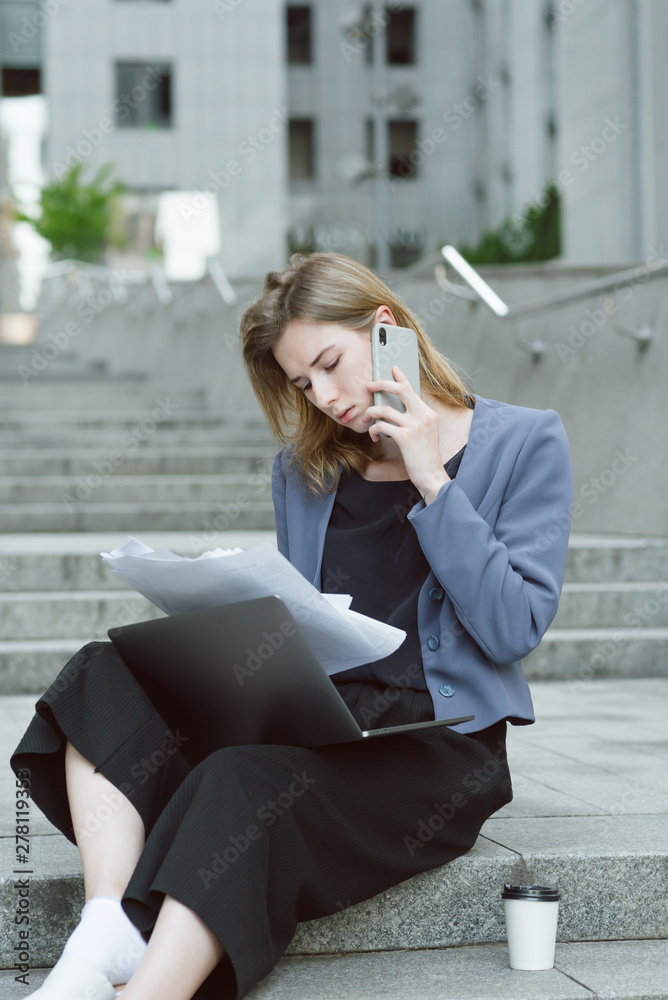 Portrait of thoughtful caucasian female office worker checking documents talking via phone. Busy employer sitting on stairs near business center with the papers, laptop and having phone conversation.
