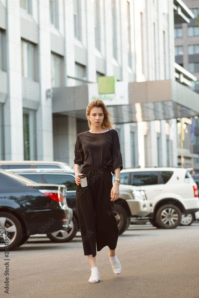 Portrait of serious young woman walking casually on the street near the business center and car parking holding take away coffee cup. Relaxed attractive girl strolling around in the urban scenery. Portrait of serious young woman walking casually on the street near the business center and car parking holding take away coffee cup. Relaxed attractive girl strolling around in the urban scenery.