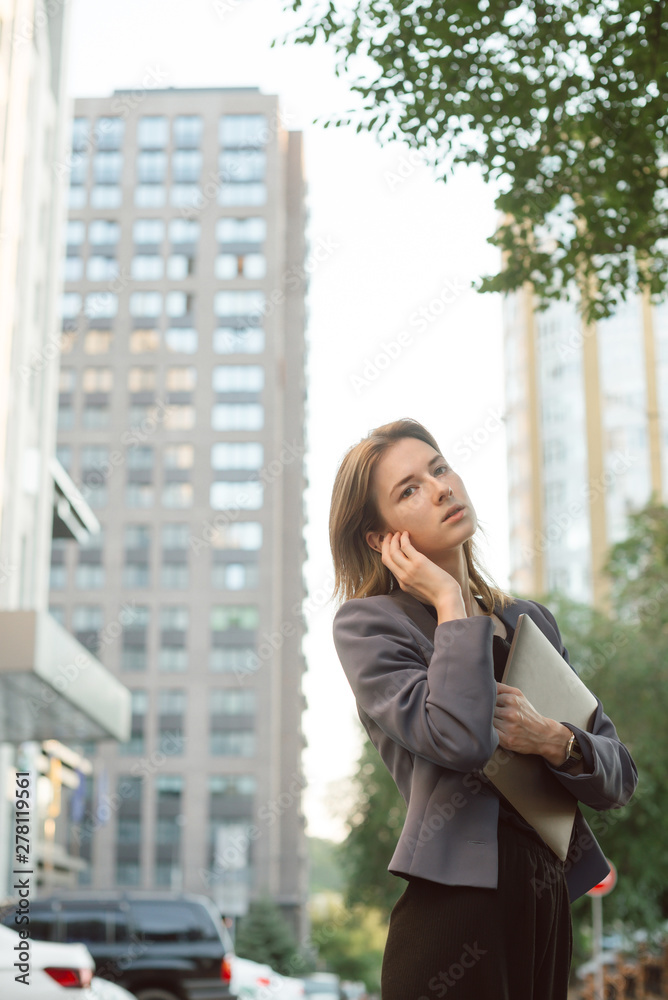 Fototapeta premium Young successful businesswoman in smart casual clothes in front of office buildings and skyscrapers. Woman holding modern laptop computer, listening to music in wireless headphones on street outdoors.