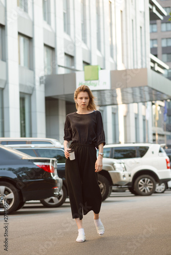 Portrait of serious young woman walking casually on the street near the business center and car parking holding take away coffee cup. Relaxed attractive girl strolling around in the urban scenery.