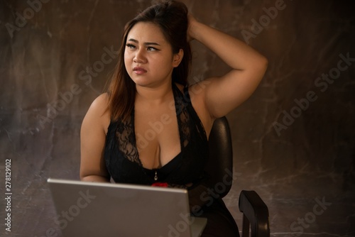 Asian plus size sexy , fat and overweight long hair model female in black dresses on brown studio background pose sitting use laptop.