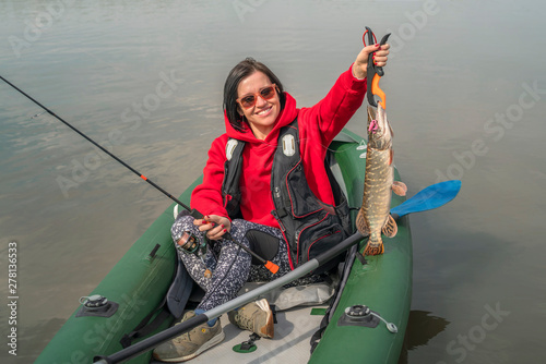 Kayak fishing. Fisher girl holding pike fish trophy on inflatable boat with fishing tackle at lake.