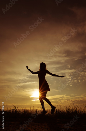 Silhouette of a girl in a dress on the background of the sunset sky and the sun, below - the grass. She is dancing. The concept of freedom, happiness.