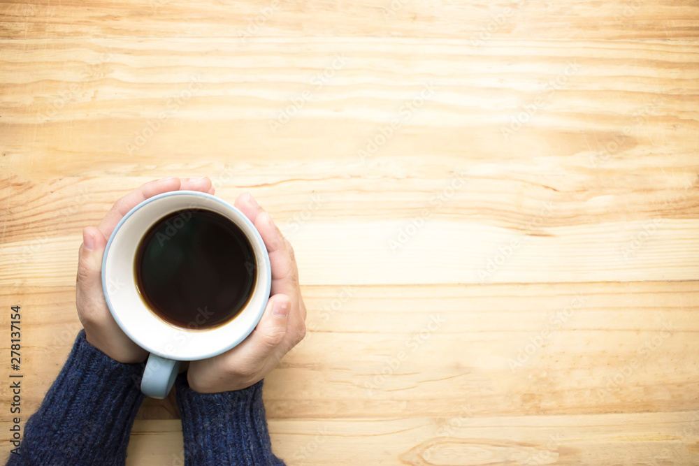 A woman wearing a blue jersey holding a cup of coffee with her hands. Place it on the wood table on the left hand side of the picture. Top view. - obrazy, fototapety, plakaty 